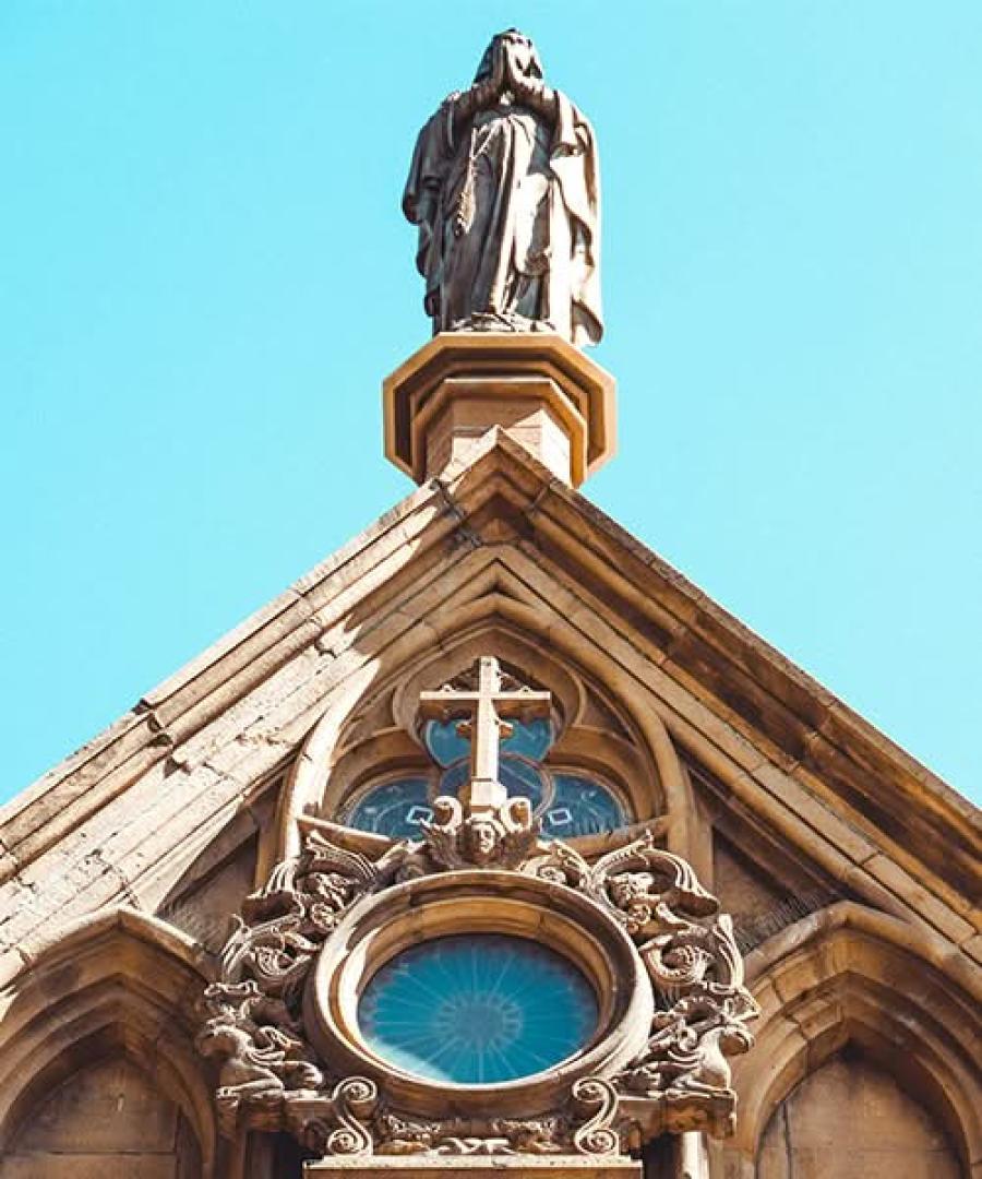 Gothic revival church facade detail showing a rose window and rooftop statue against a blue sky.